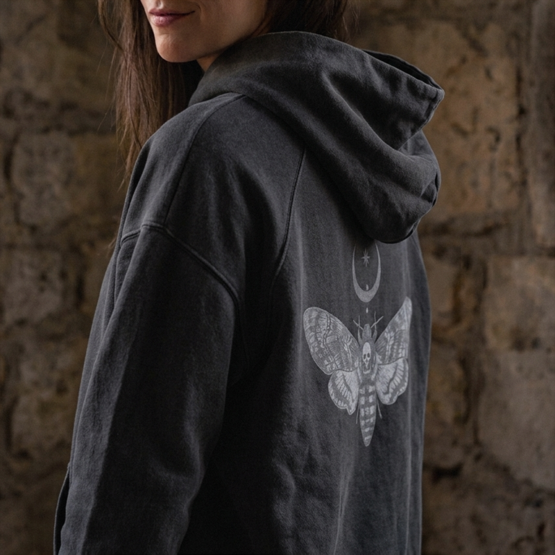 A person wearing a dark charcoal gray hooded sweatshirt with a large white moth and crescent moon graphic printed on the back, posed against a rustic stone wall.