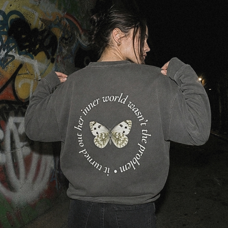 A person viewed from behind wearing a dark gray sweatshirt with a circular graphic on the back featuring a butterfly and the text 'it turned out her inner world wasn't the problem', set against a dark outdoor background with graffiti.