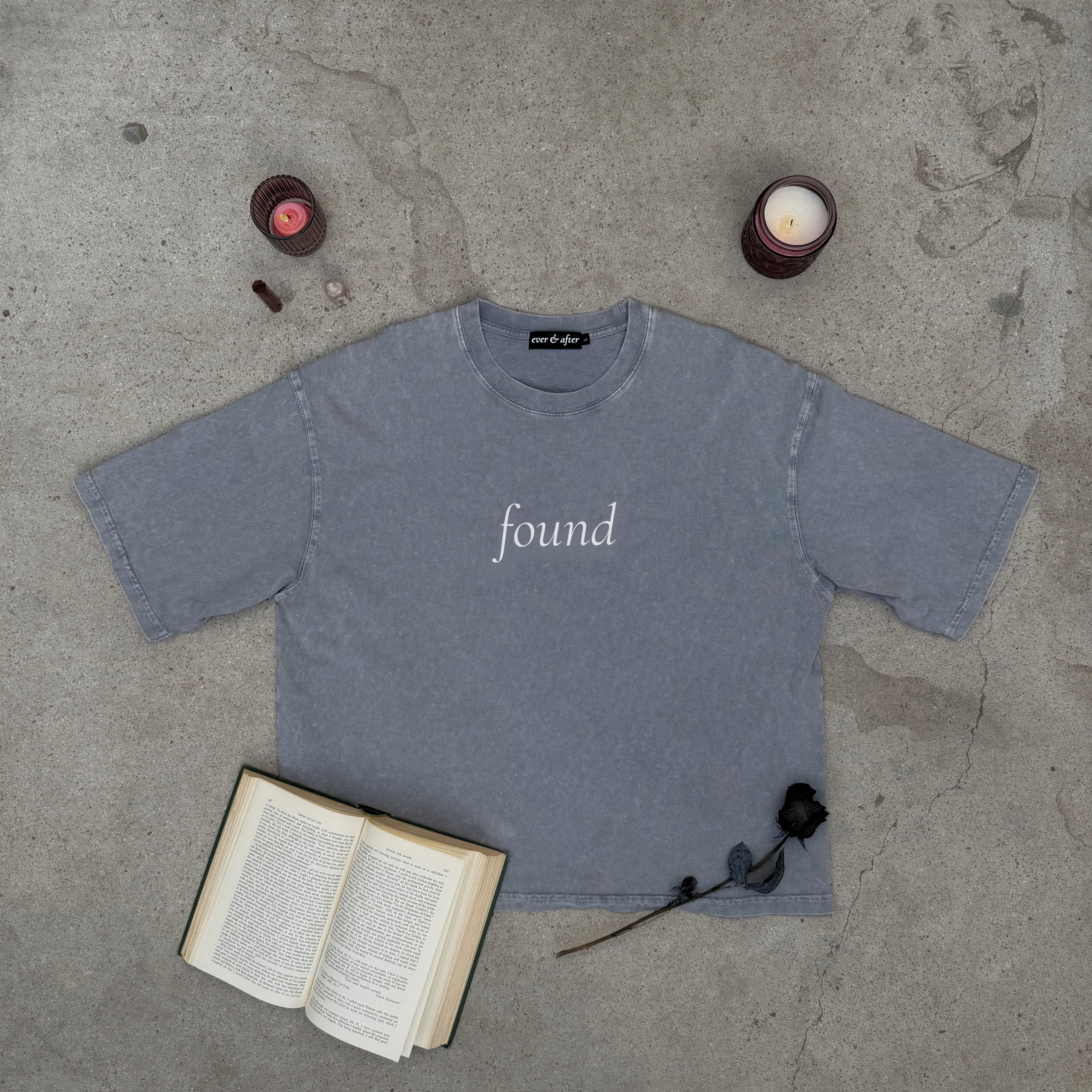 light gray boxy tee layflat on concrete with rose, candles, crystals, and an old book  text found on center chest