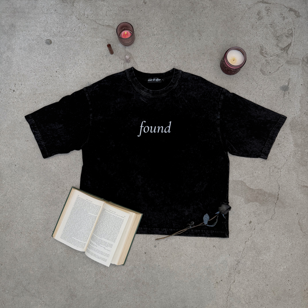 faded black boxy tee layflat on concrete with rose, candles, crystals, and an old book  text found on center chest