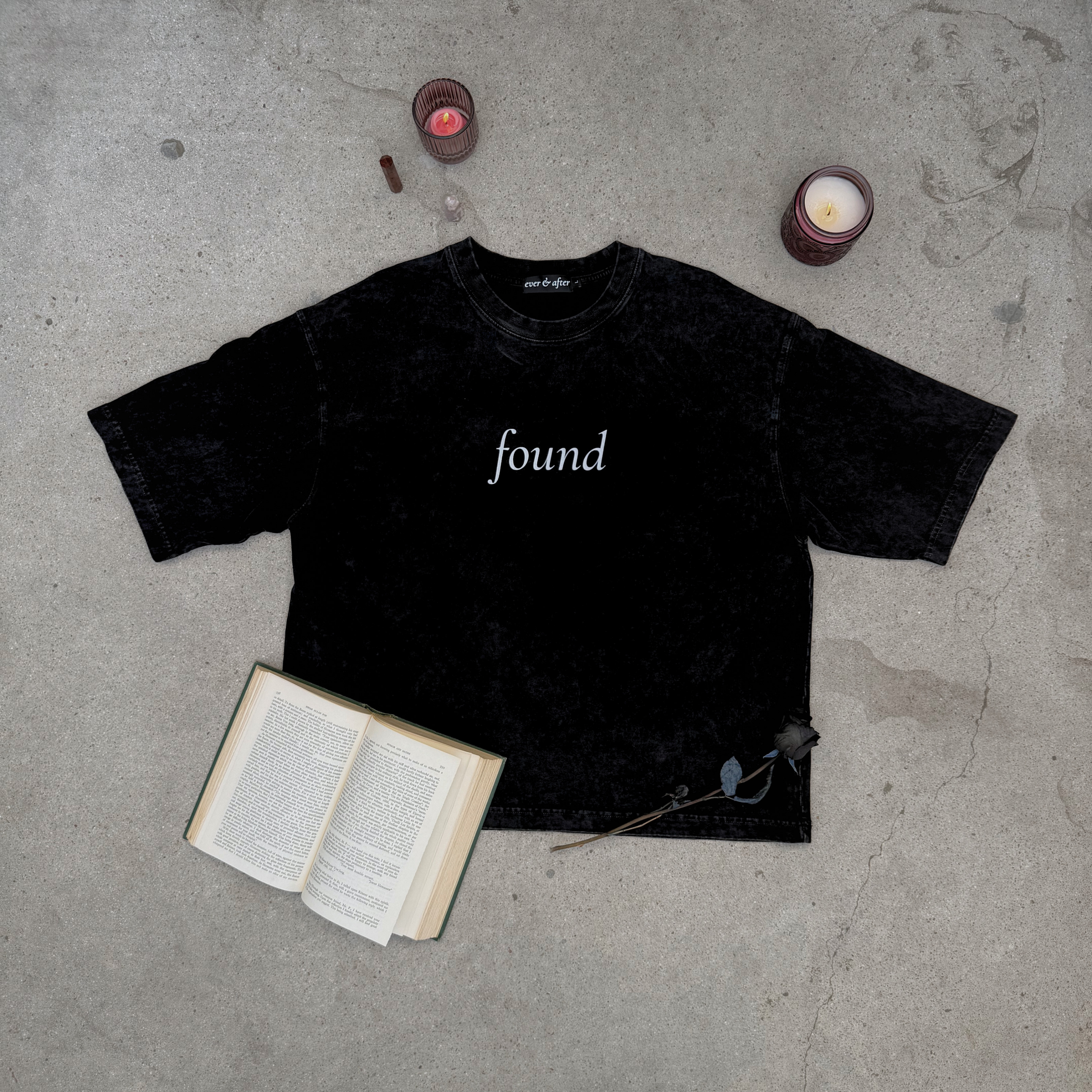 faded black boxy tee layflat on concrete with rose, candles, crystals, and an old book  text found on center chest