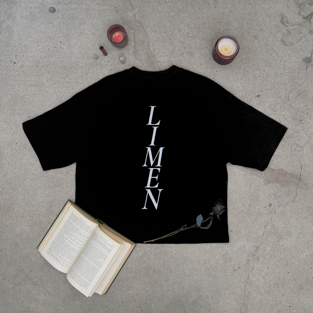 back of distressed black boxy tee with word LIMEN vertically down the center of the tee layflat on concrete with rose, candles, crystals, and an old book  