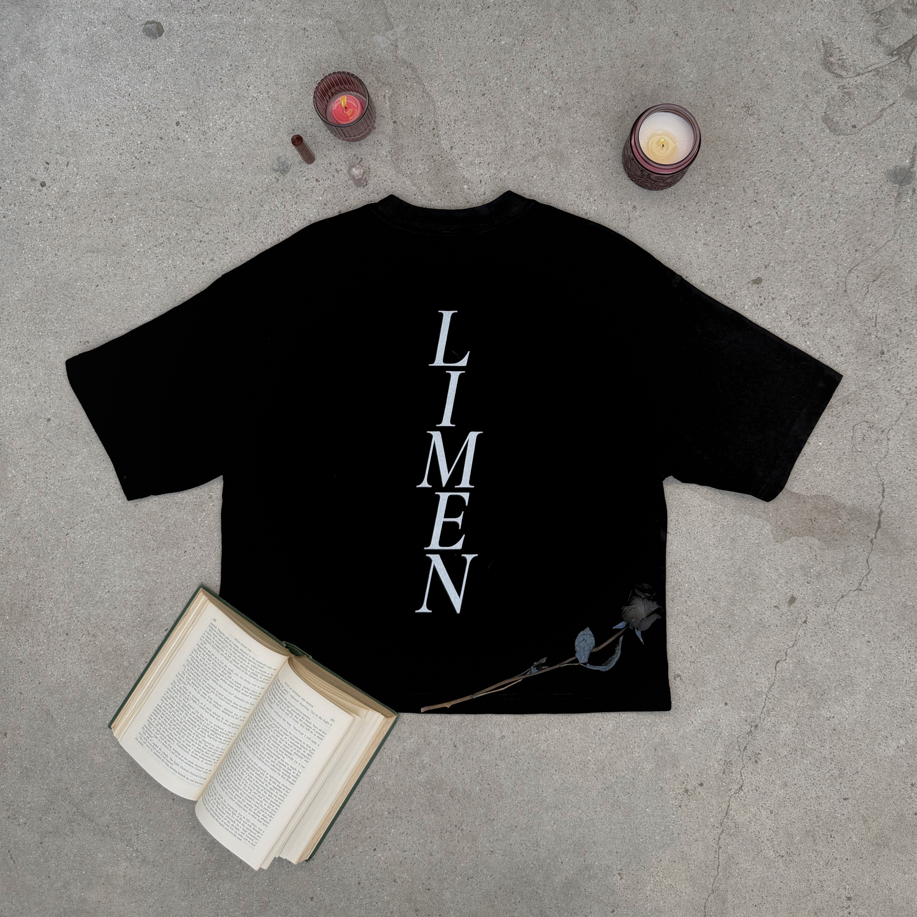 back of distressed black boxy tee with word LIMEN vertically down the center of the tee layflat on concrete with rose, candles, crystals, and an old book  