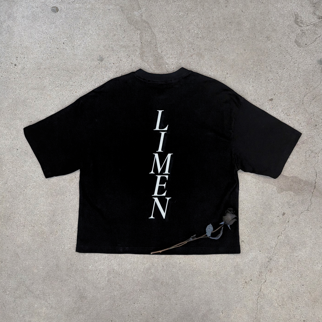 laying on concrete with rose the back of distressed black boxy tee with word LIMEN vertically down the center of the tee 
