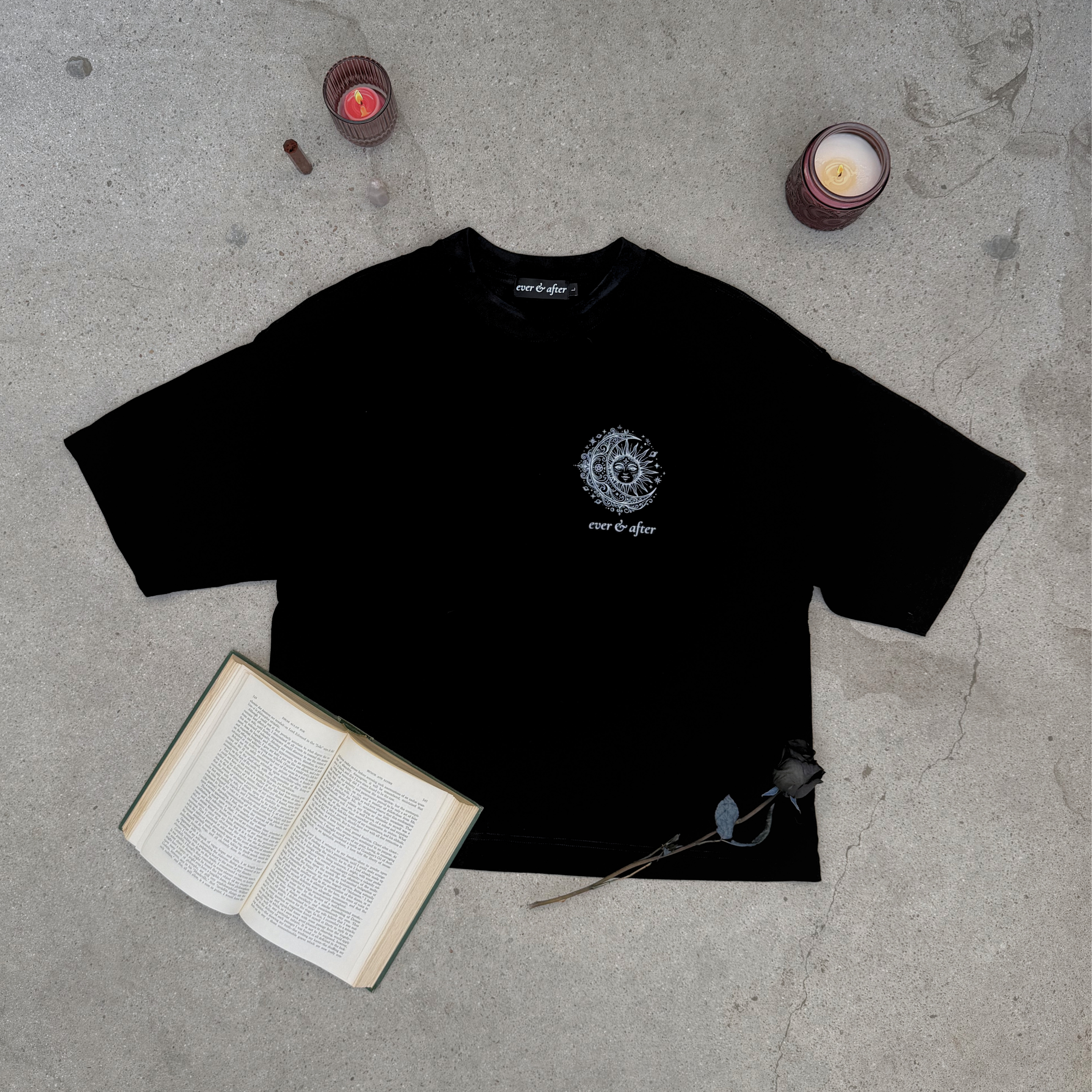 distressed black boxy tee with sun and moon design small on left chest with small brand word mark: ever & after layflat on concrete with rose, candles, crystals, and an old book 