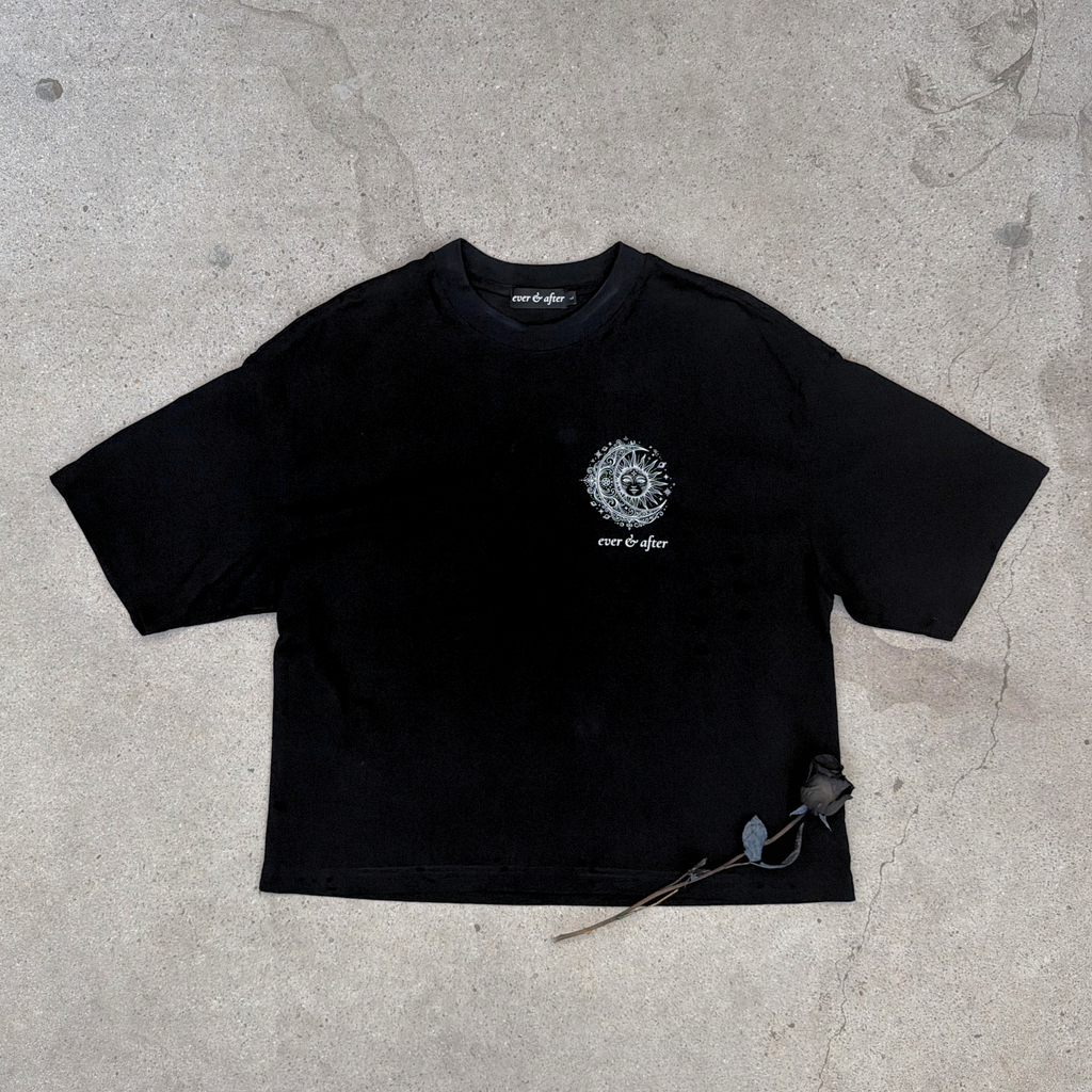 distressed black boxy tee with sun and moon design small on left chest with small brand word mark: ever & after layflat on concrete with rose 