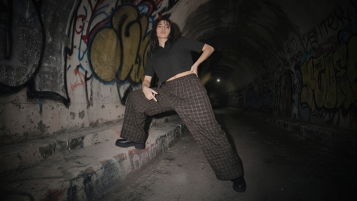 A person posing in a dark, graffiti-covered tunnel, wearing a black short-sleeve crop top and wide-leg brown plaid trousers.