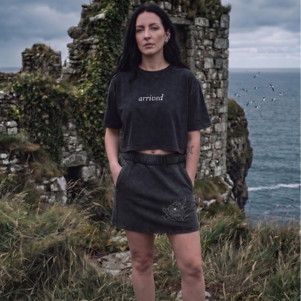Woman wearing a black t-shirt with 'arrived' text, standing in front of stone ruins by the sea.