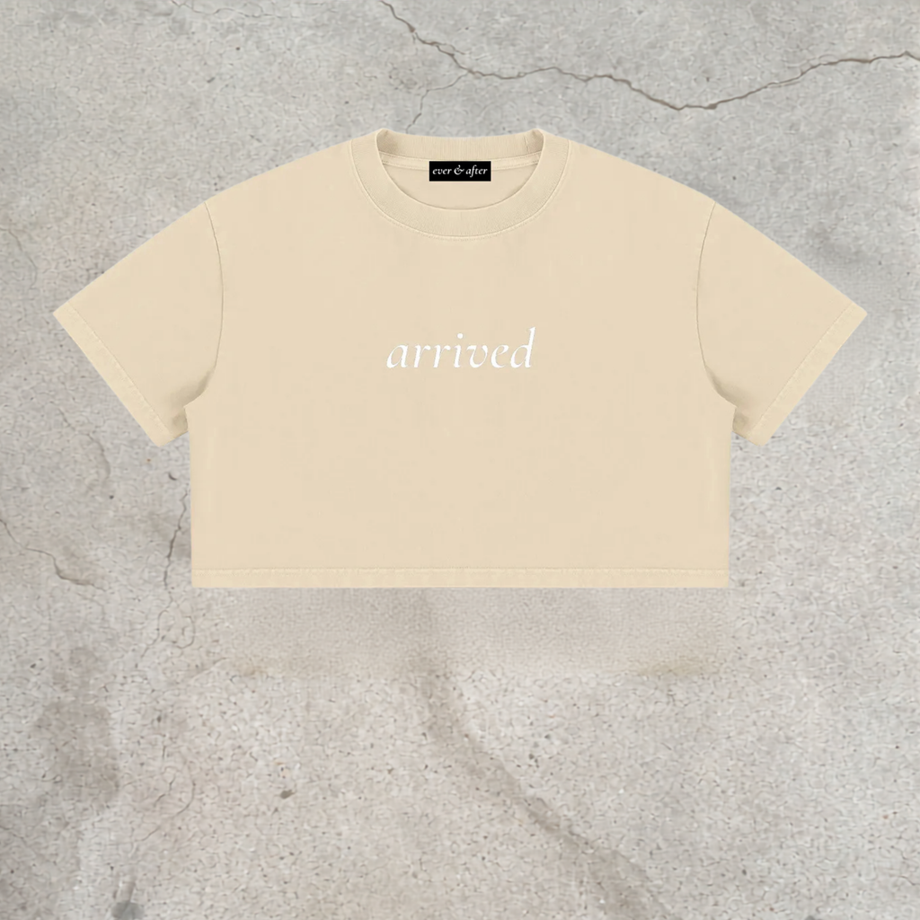 A cream-colored short-sleeve crop top with the word 'arrived' printed in white script across the chest, laid flat on a textured gray concrete surface.