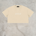 A cream-colored short-sleeve crop top with the word 'arrived' printed in white script across the chest, laid flat on a textured gray concrete surface.