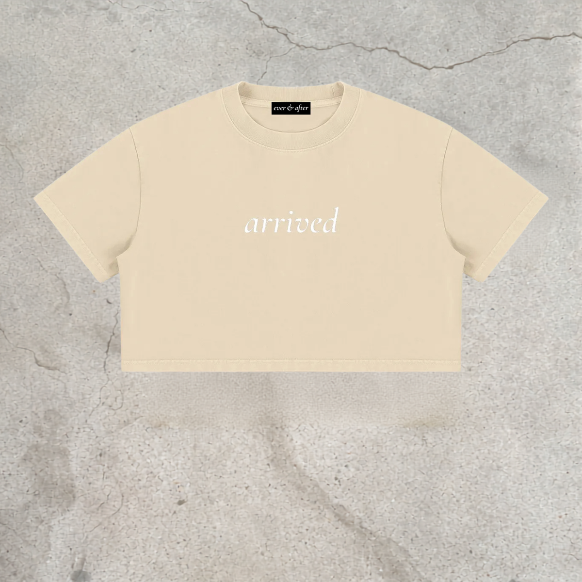 A cream-colored short-sleeve crop top with the word 'arrived' printed in white script across the chest, laid flat on a textured gray concrete surface.