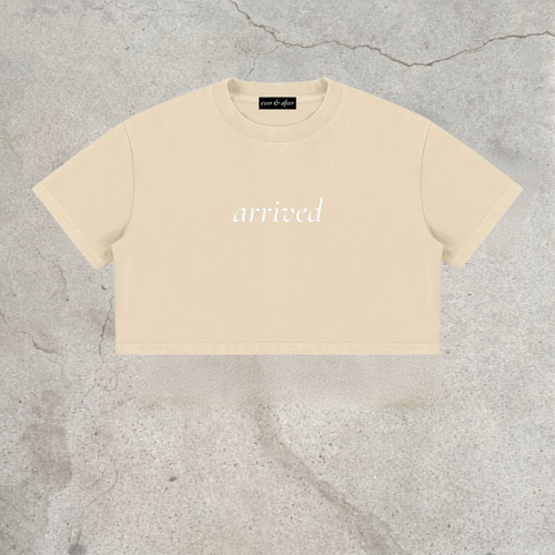 A cream-colored short-sleeve crop top with the word 'arrived' printed in white script across the chest, laid flat on a textured gray concrete surface.