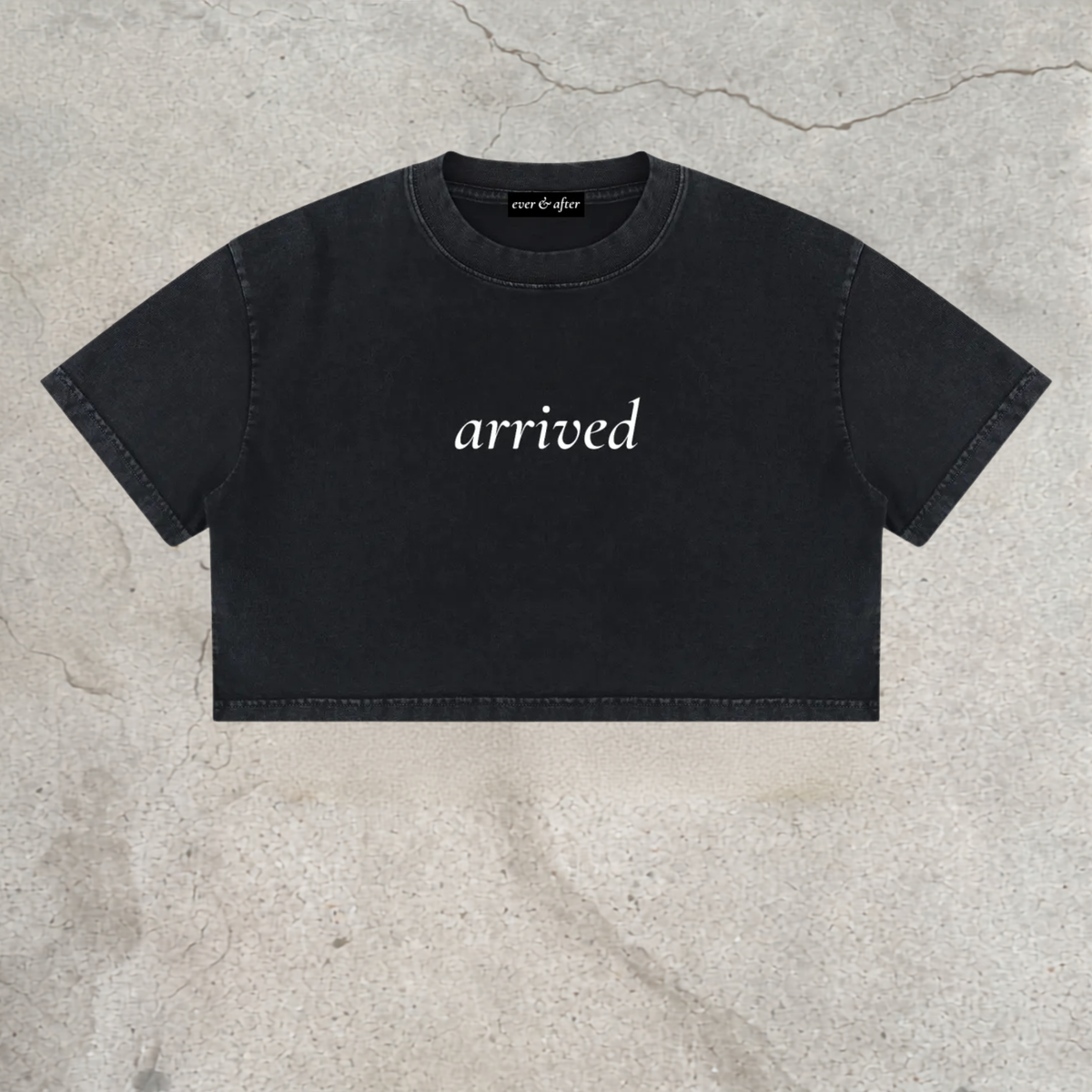 A black cropped t-shirt with the word 'arrived' printed in white script across the chest, laid flat on a textured concrete surface.