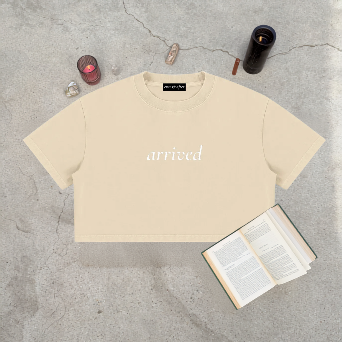A cream-colored cropped t-shirt with the word 'arrived' printed in white script on the chest, styled in a flat-lay on a gray concrete surface with an open book, crystals, and candles.