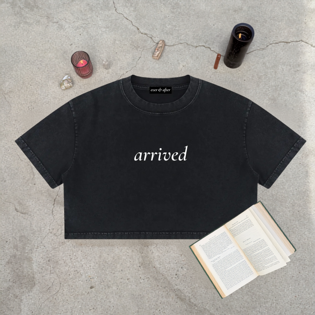 A black cropped t-shirt with the word 'arrived' printed in white script across the chest, styled in a flat-lay on a gray concrete surface with an open book, crystals, and candles.