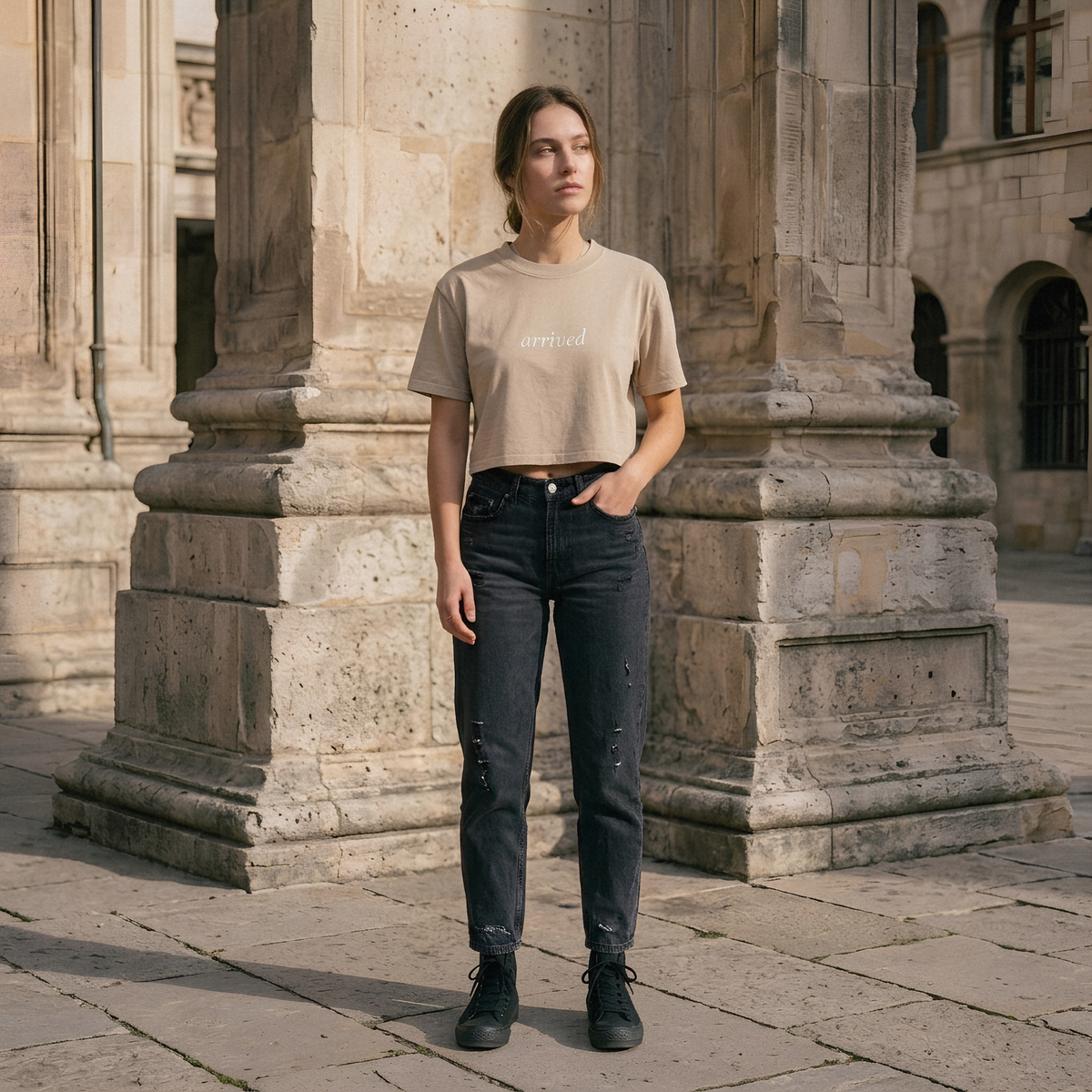 A woman stands outdoors between large stone columns, wearing a beige cropped T-shirt with the word 'affixed' printed across the chest, dark distressed jeans, and black shoes.