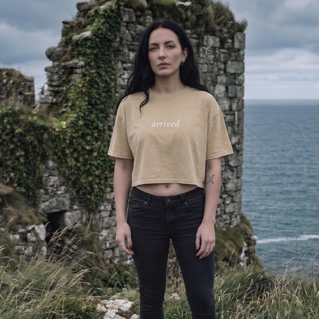 Woman wearing a beige t-shirt with 'arrived' text, standing in front of stone ruins and ocean.