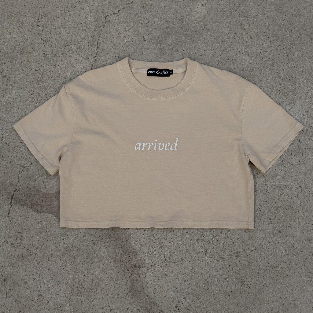 Beige t-shirt with 'arrived' text on a gray concrete surface