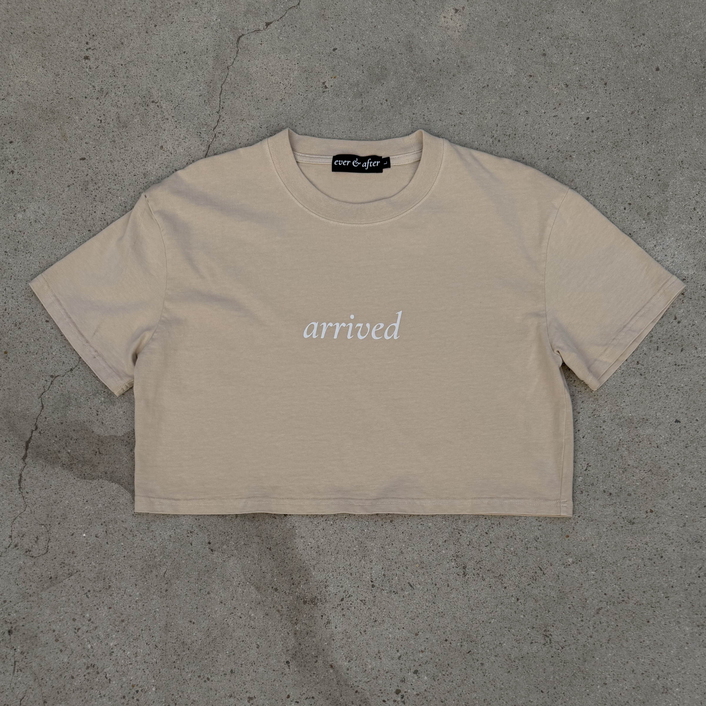 Beige t-shirt with 'arrived' text on a gray concrete surface