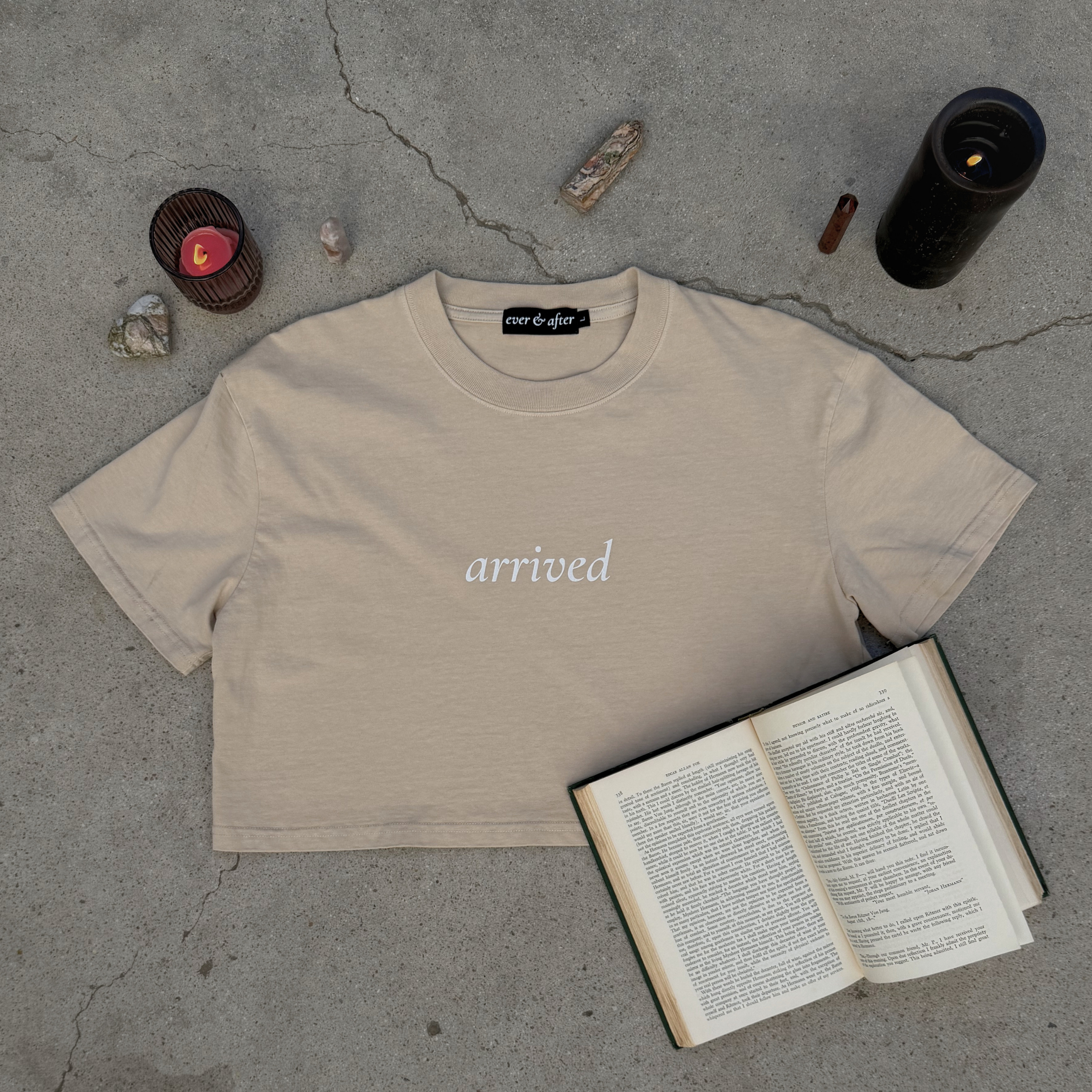 Beige t-shirt with 'arrived' text on a concrete surface with candles and an open book.