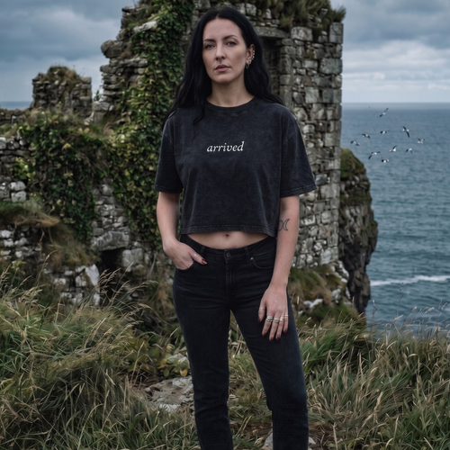 Person wearing a black t-shirt with 'arrived' text, standing in front of stone ruins and ocean.