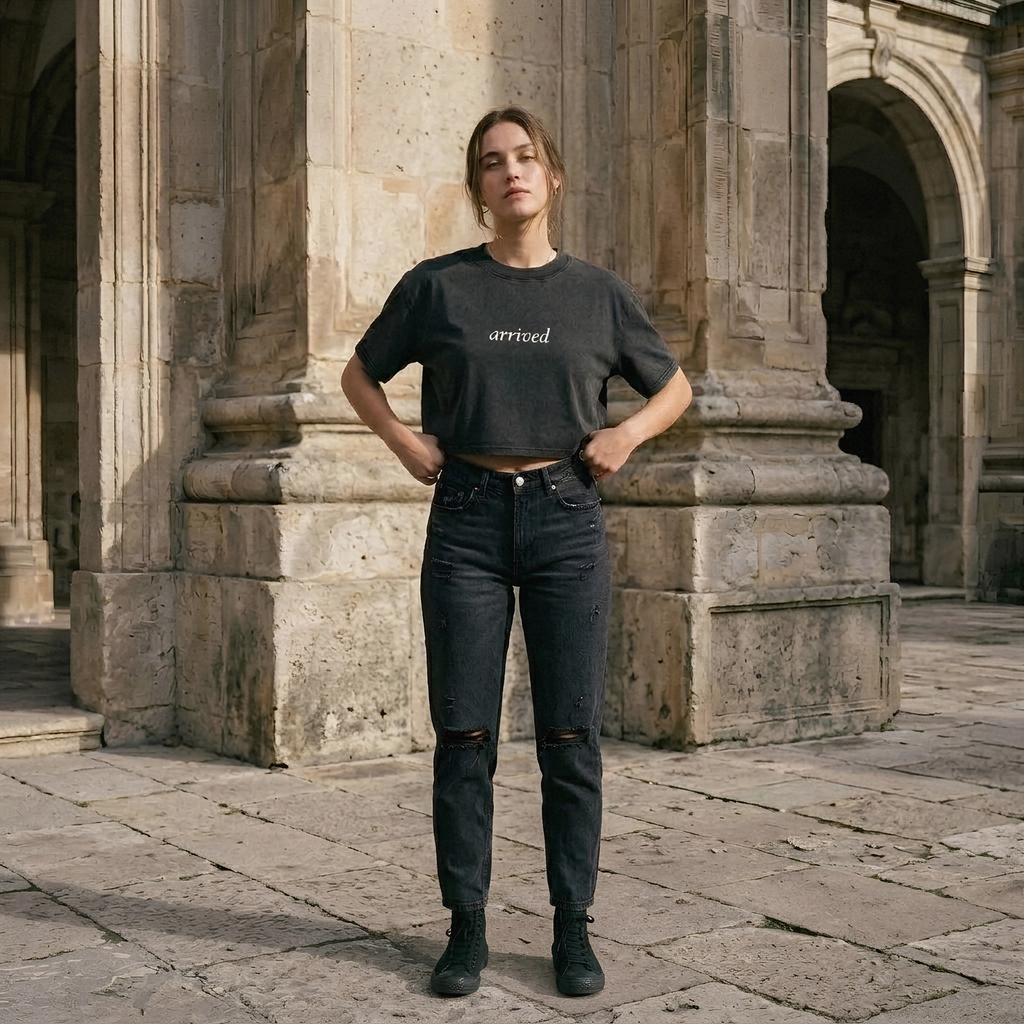 A woman stands outdoors in front of a stone archway wearing a dark charcoal cropped T-shirt with the word 'arrived' printed in white across the chest, dark distressed jeans, and black ankle boots.
