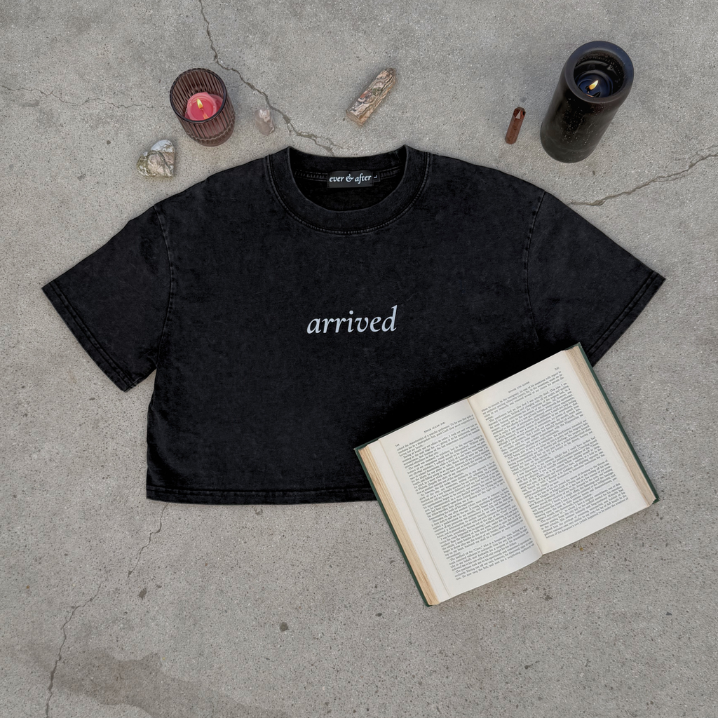 Black t-shirt with 'arrived' text on a concrete surface with an open book and candles.