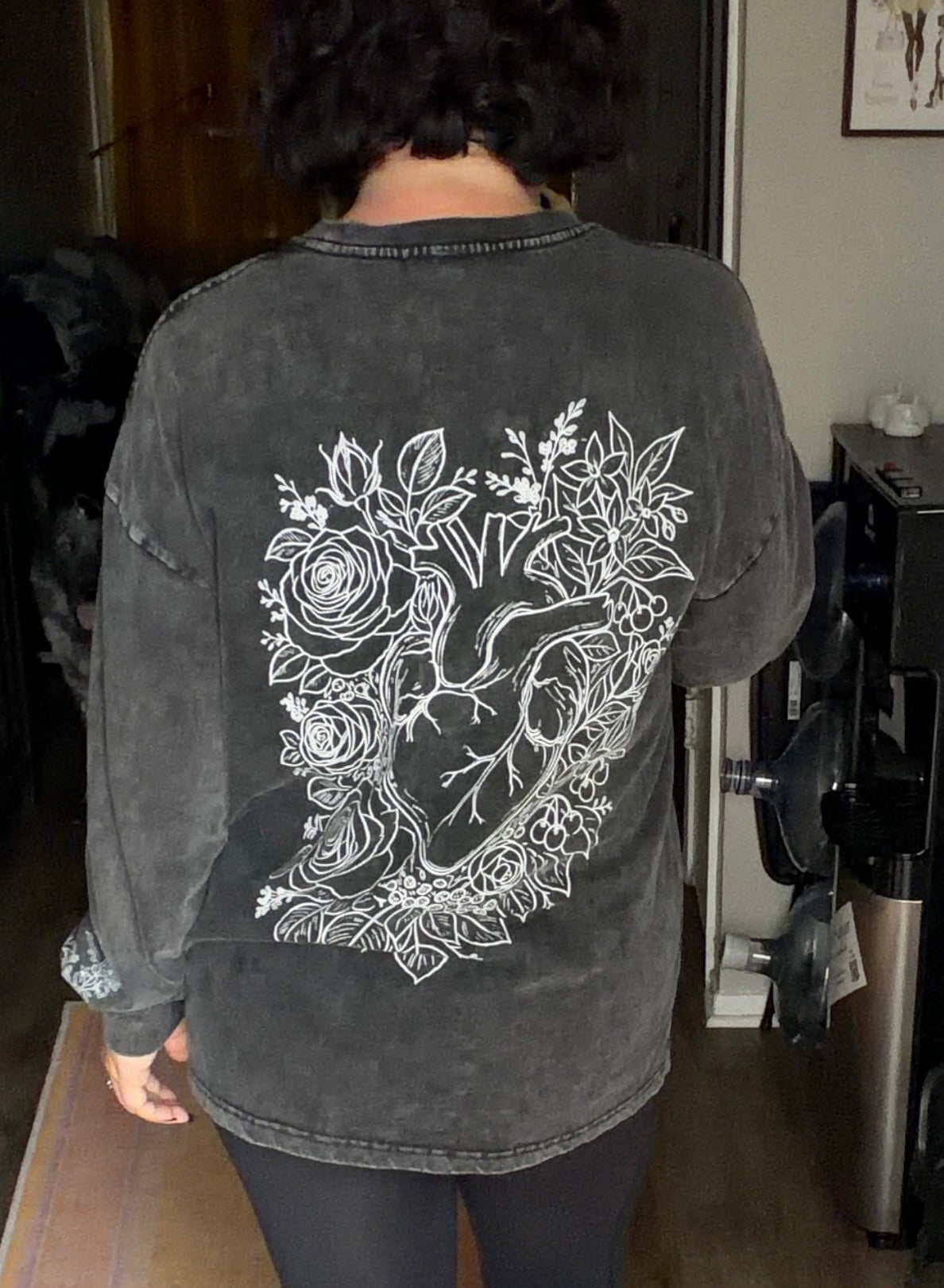 A person viewed from behind wearing a faded black graphic sweatshirt with a large white anatomical heart and floral design on the back, paired with black leggings and black-and-white sneakers in an indoor room.