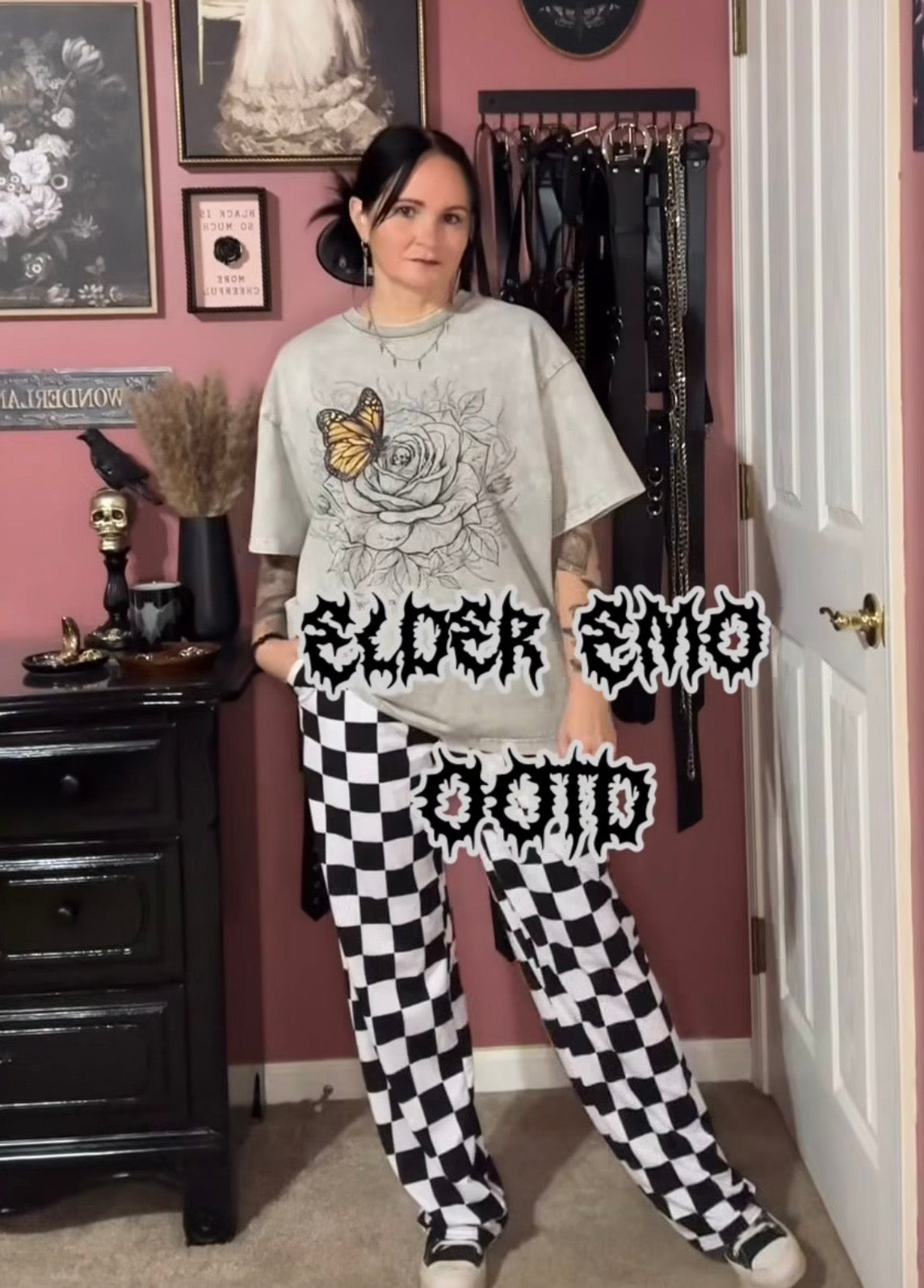 A woman stands in a room wearing a light gray graphic t-shirt with a black and yellow rose design and black-and-white checkered pants. She is posed in front of a pink wall with a black clothing rack and a dark wooden dresser.