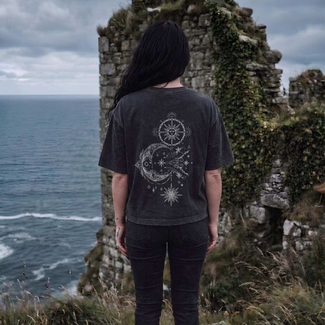 Person wearing a dark t-shirt with a celestial design, standing on a cliff overlooking the ocean.
