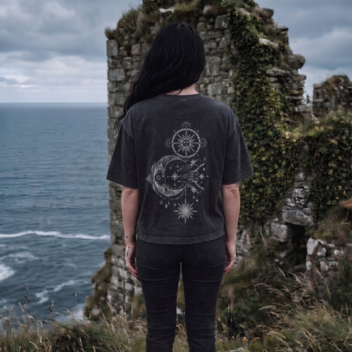 Person wearing a dark t-shirt with a celestial design, standing on a cliff overlooking the ocean.