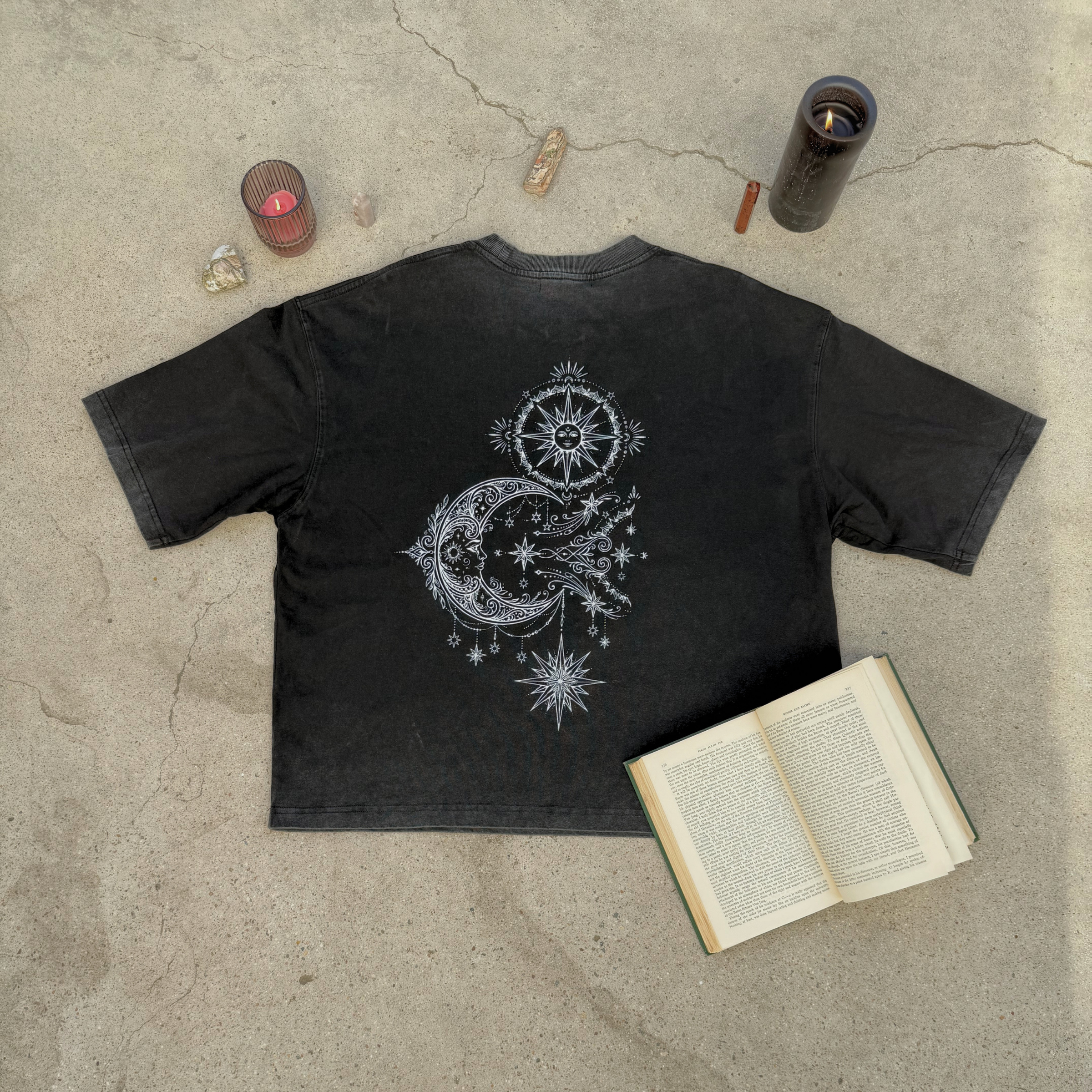 Black t-shirt with celestial design on a concrete surface with candles and an open book.