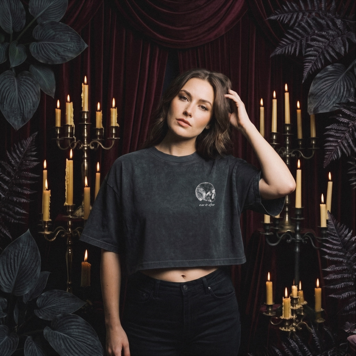 A woman stands in a dark, gothic-themed room wearing a washed black cropped t-shirt with a small white graphic on the left chest, paired with black jeans. The background features deep red velvet drapery, lit candles on brass candelabras, and dark foliage.