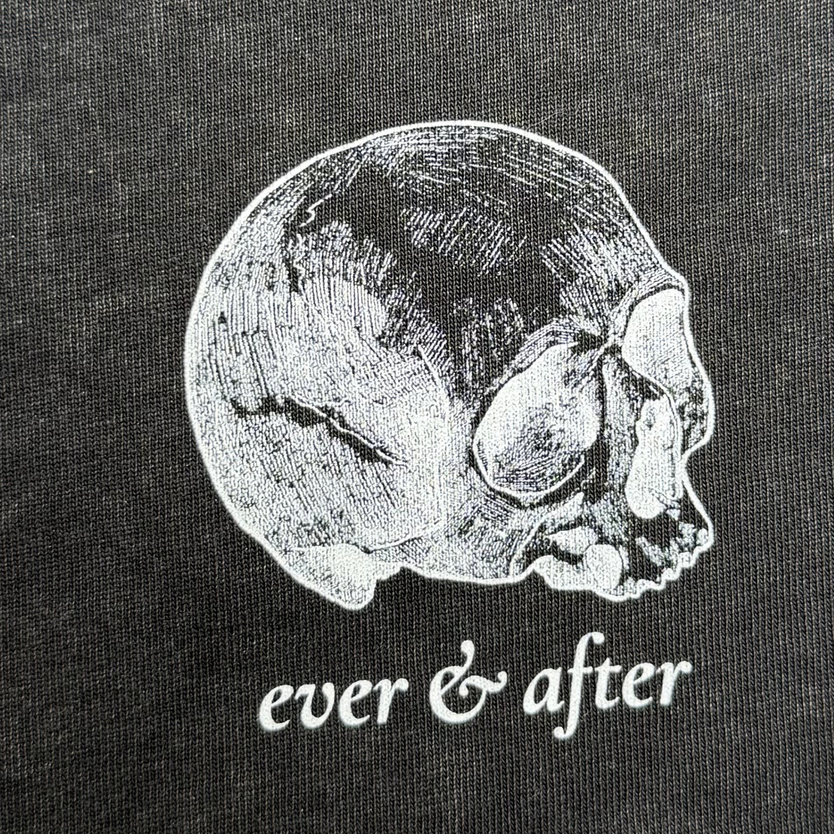 Close-up of a black fabric surface featuring a white line-art skull graphic and the text 'ever & after' in white script below it.