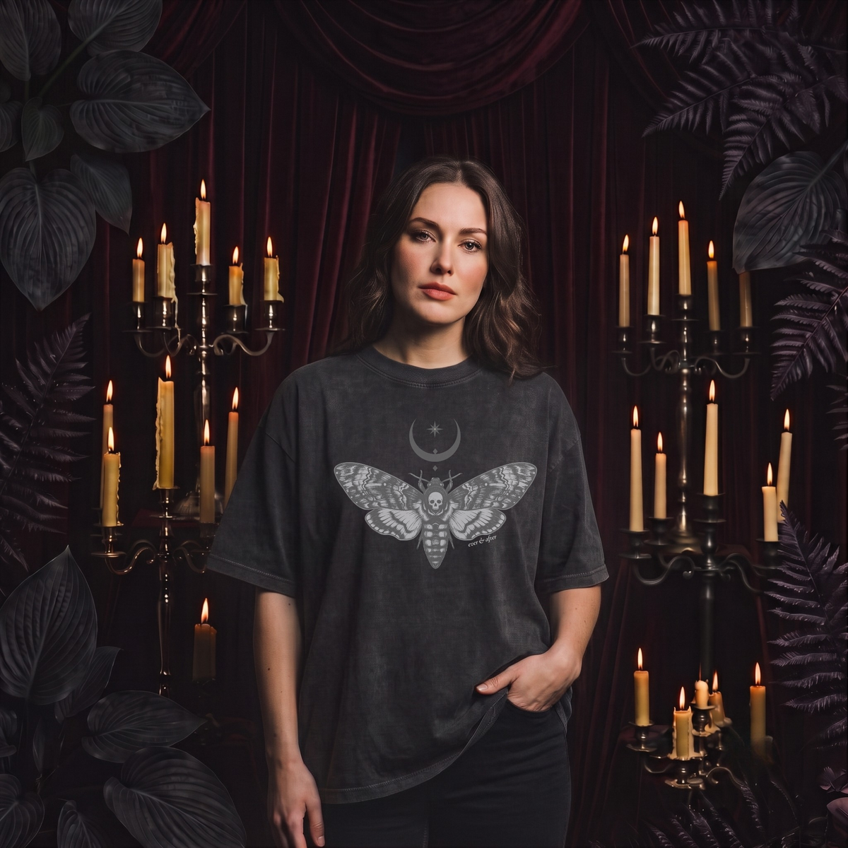 A woman stands in a dark, moody setting with red velvet drapery and lit candles, wearing a faded charcoal gray t-shirt featuring a detailed gray moth and crescent moon graphic on the front.