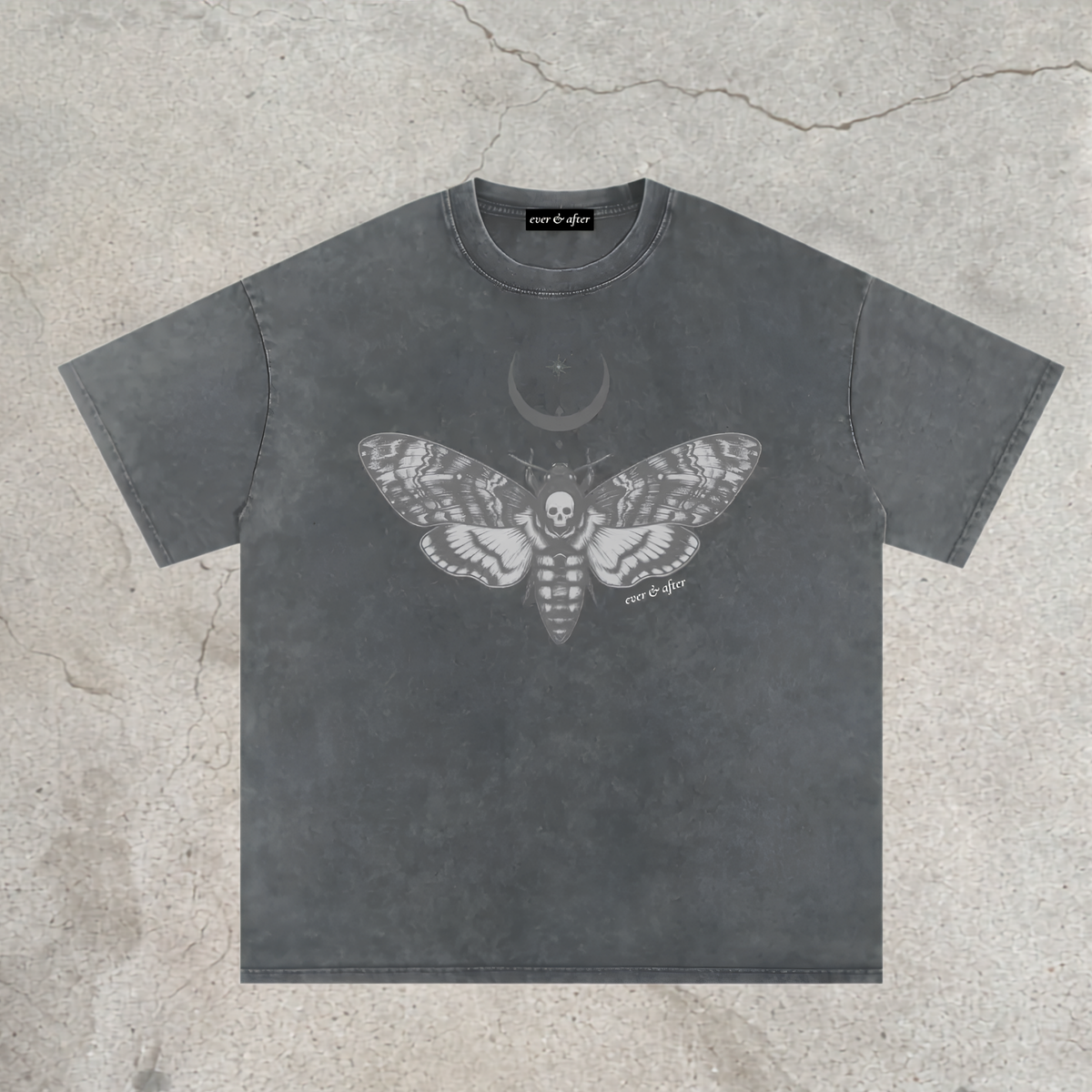 A dark gray acid-wash t-shirt with a detailed white graphic of a moth, a crescent moon, and a skull on the front, laid flat on a textured concrete background.