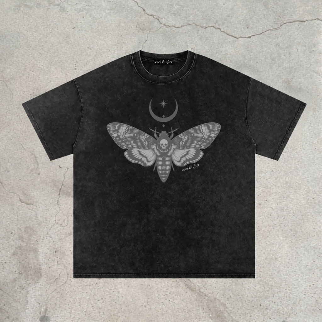 A black acid-wash t-shirt with a gray graphic of a moth containing a skull, a crescent moon, and stars on the chest. The t-shirt is laid flat on a textured concrete background.