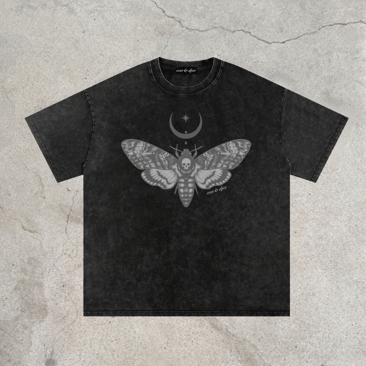 A black acid-wash t-shirt with a gray graphic of a moth containing a skull, a crescent moon, and stars on the chest. The t-shirt is laid flat on a textured concrete background.