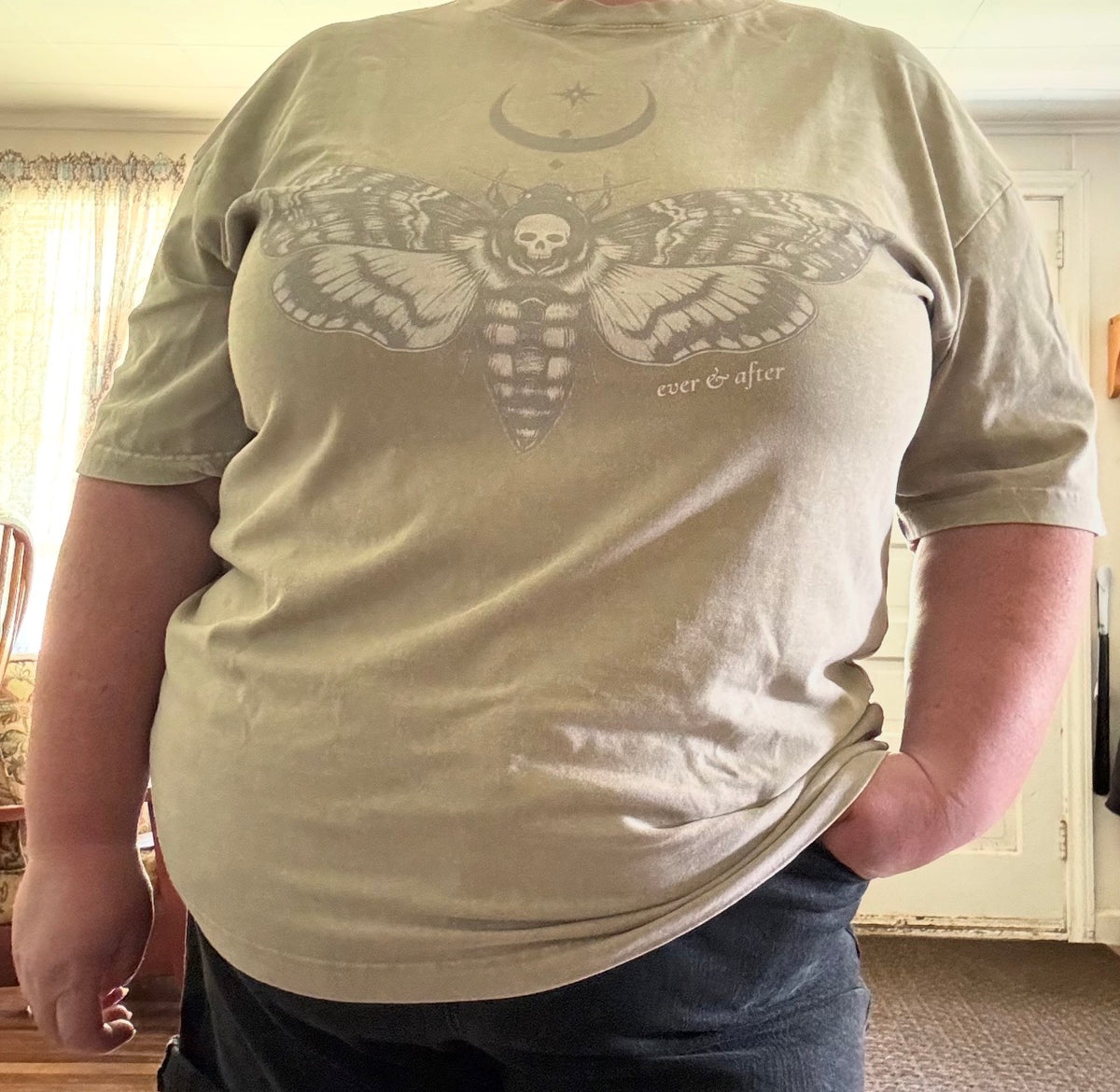 A person wearing a light beige graphic t-shirt with a large moth and skull design on the front, standing in a home interior.