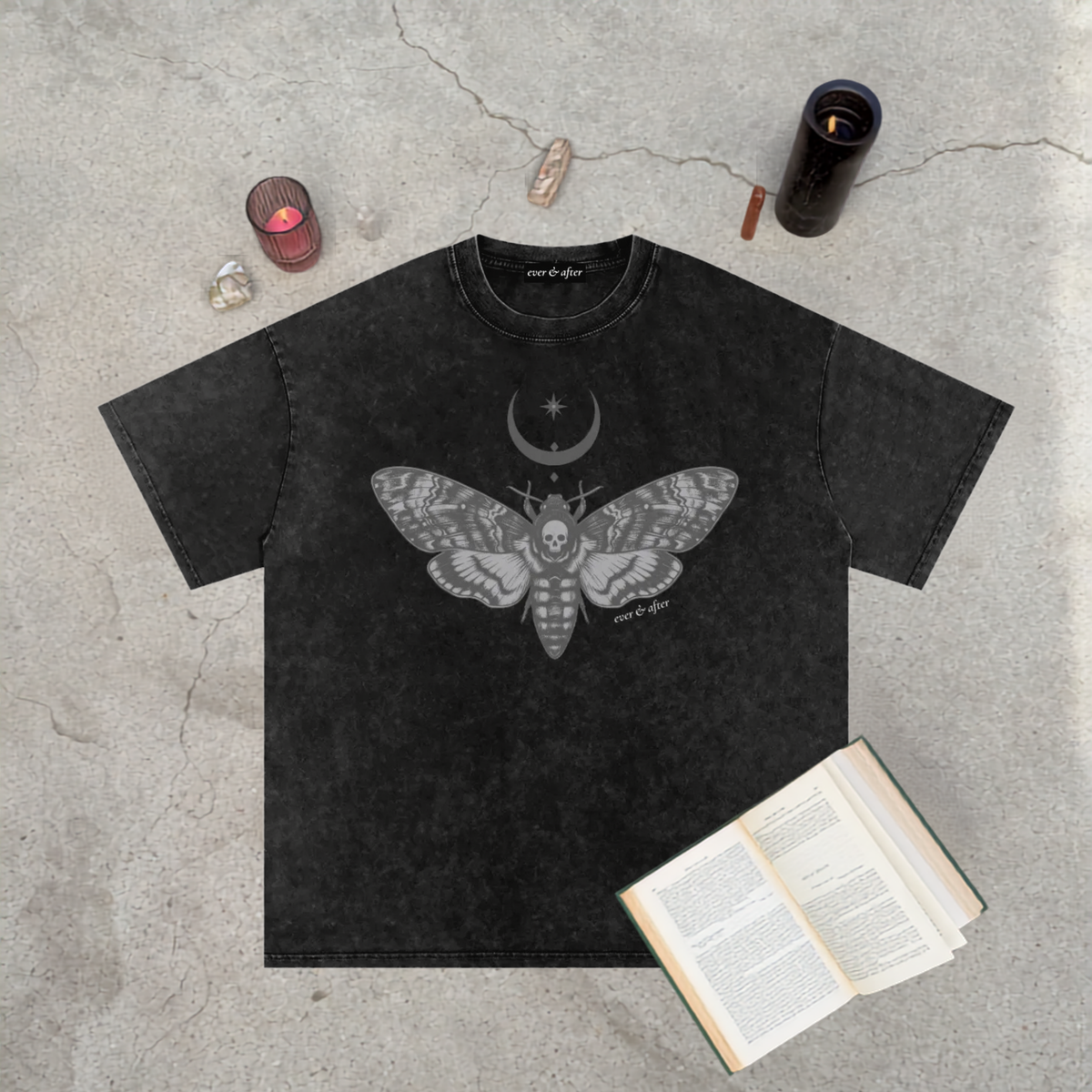 A black acid-wash graphic t-shirt featuring a detailed gray moth and crescent moon design, styled in a flat-lay on a textured concrete surface with a book, crystals, and candles.