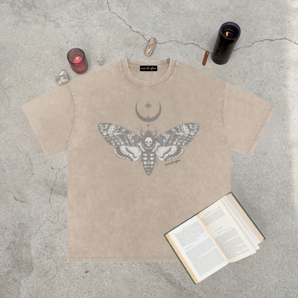 A beige graphic t-shirt featuring a gray moth, crescent moon, and star design, styled in a flat-lay on a concrete surface with a book, crystals, and candles.
