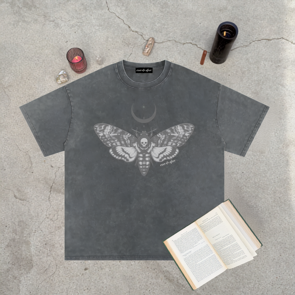 A dark gray washed t-shirt with a detailed gray moth and crescent moon graphic, laid flat on a concrete surface with occult-themed props including a book, crystals, and candles.