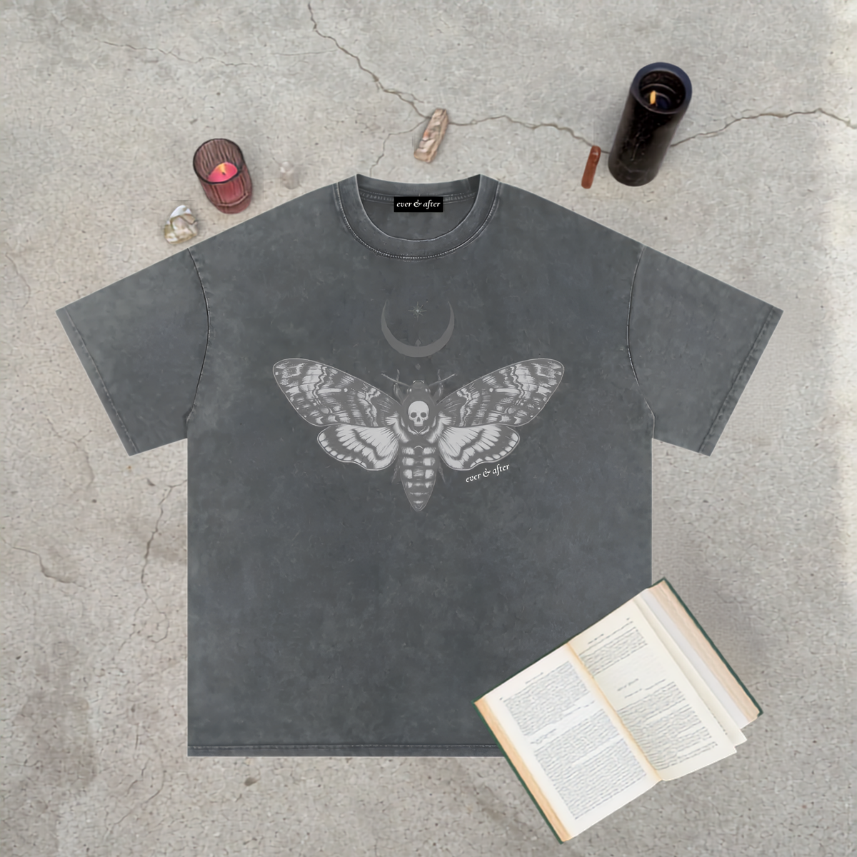 A dark gray washed t-shirt with a detailed gray moth and crescent moon graphic, laid flat on a concrete surface with occult-themed props including a book, crystals, and candles.