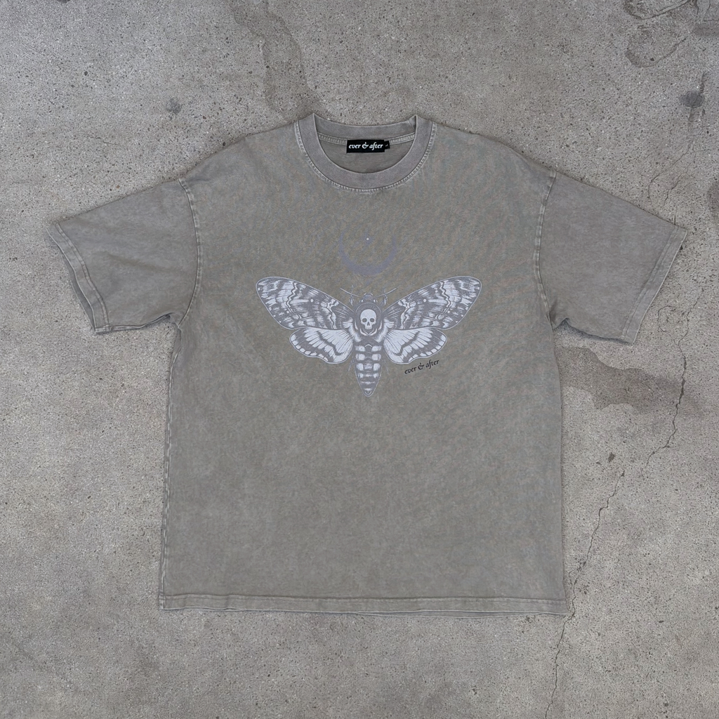 A light khaki t-shirt featuring a large white graphic of a moth with a skull and crossbones inside, laid flat on a textured concrete surface.