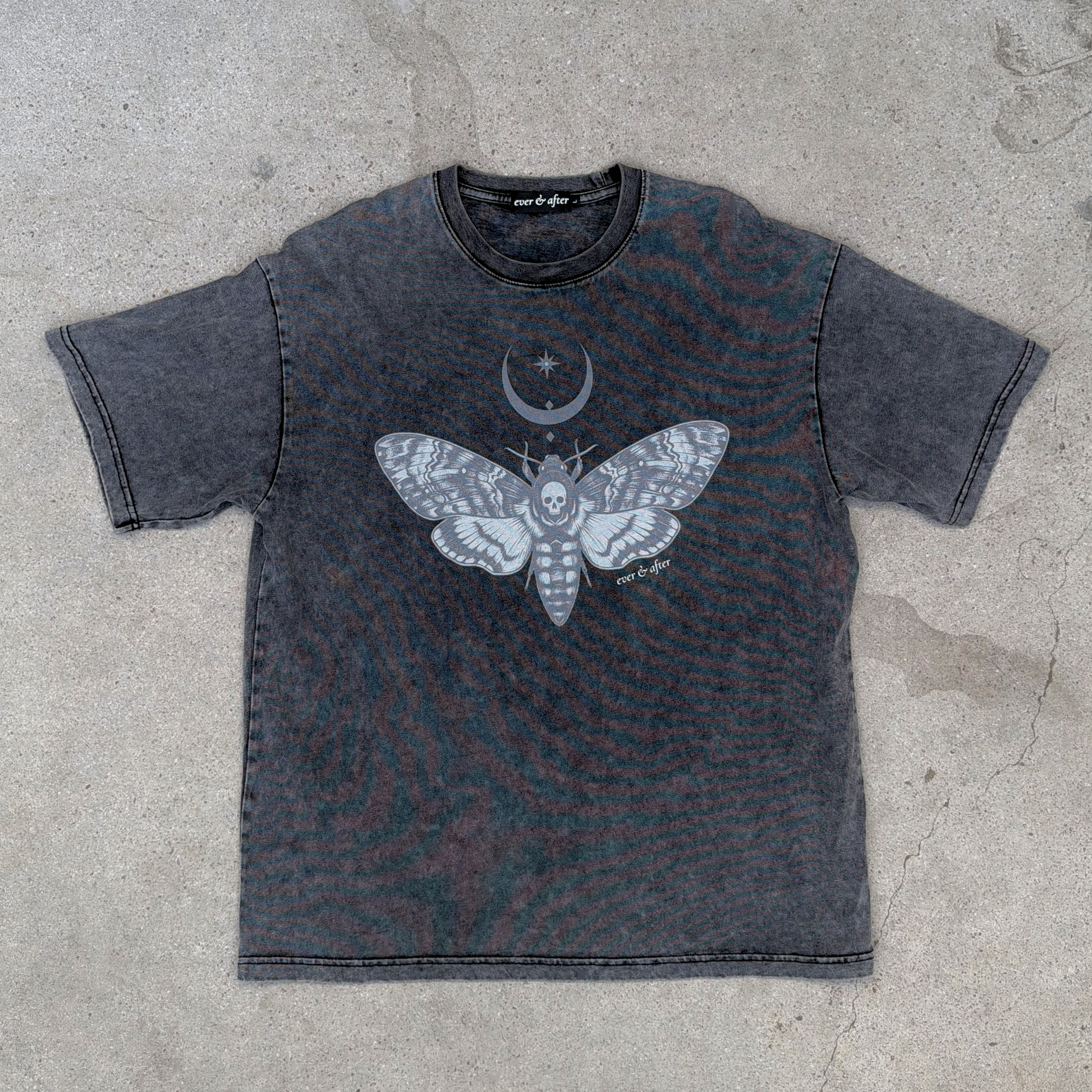A washed charcoal gray t-shirt with a large white and gray moth graphic and a crescent moon with a star on the chest, laid flat on a concrete surface.