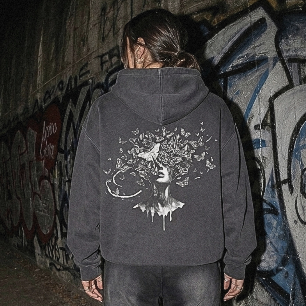 A person viewed from behind wearing a dark gray hoodie with a large white graphic print on the back, paired with dark sweatpants and white sneakers, standing in front of a graffiti-covered wall.