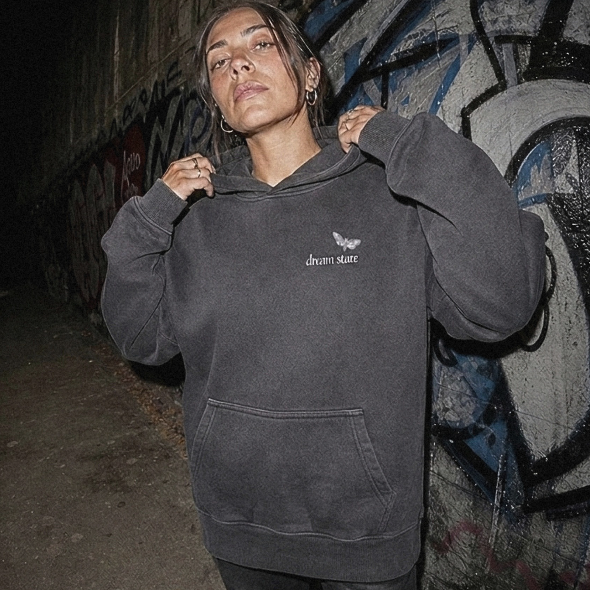 A person wearing a dark charcoal gray hooded sweatshirt with a small white graphic on the chest, standing in a dimly lit urban alley with graffiti-covered walls.