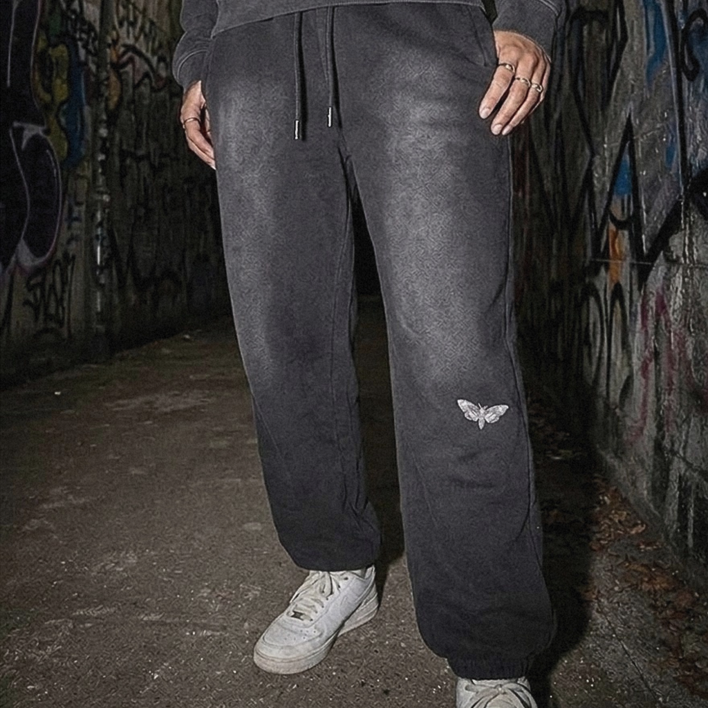 A person wearing a dark charcoal gray matching sweatshirt and jogger pants set, standing in a graffiti-covered alleyway. The jogger pants feature a small white embroidered butterfly logo on the lower left leg, and the person is wearing white sneakers.