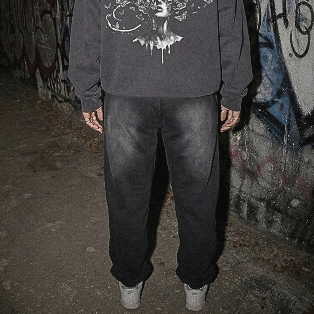 A person viewed from behind wearing a dark gray hoodie with a large white graphic print on the back, paired with dark sweatpants and white sneakers, standing in front of a graffiti-covered wall.
