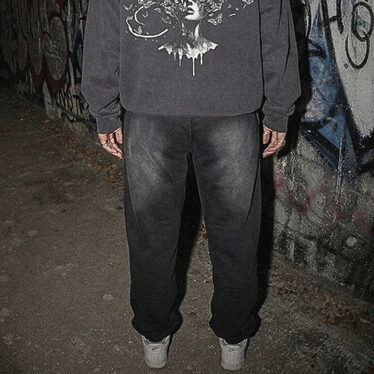 A person viewed from behind wearing a dark gray hoodie with a large white graphic print on the back, paired with dark sweatpants and white sneakers, standing in front of a graffiti-covered wall.